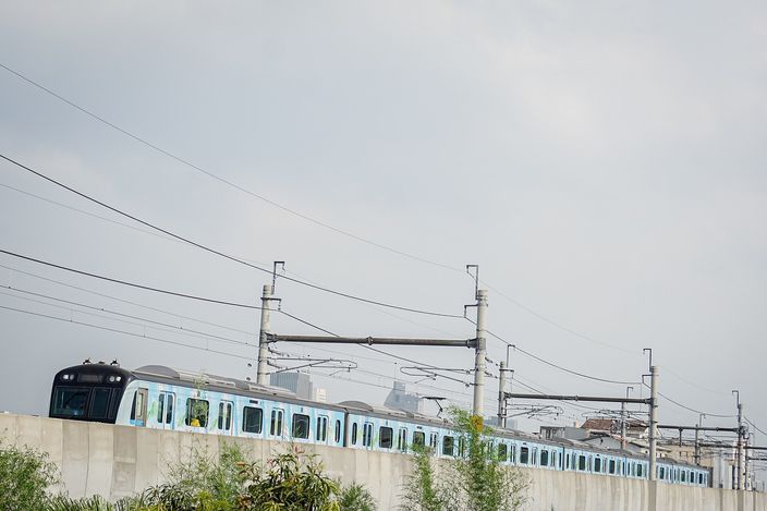 A teal-and-white MRT Jakarta train running on an elevated viaduct against a hazy sky, with overhead catenary masts and distant towers behind