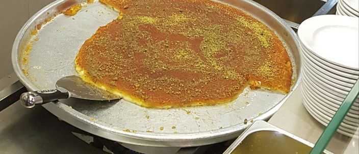A large round metal tray of knafeh on a warmer, its smooth orange top scattered with crushed pistachio, with a serving spatula mid-cut and a stack of plates and a tray of chopped pistachio beside it.