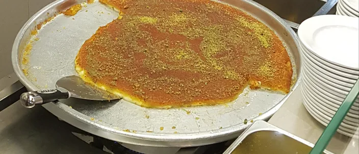 A large round metal tray of knafeh on a warmer, its smooth orange top scattered with crushed pistachio, with a serving spatula mid-cut and a stack of plates and a tray of chopped pistachio beside it.
