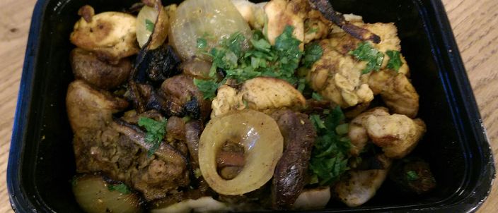 A portion of me'orav yerushalmi: griddled chicken pieces and darker offal mixed with cooked onion rings and chopped fresh herbs, served in a dark container on a wooden table.