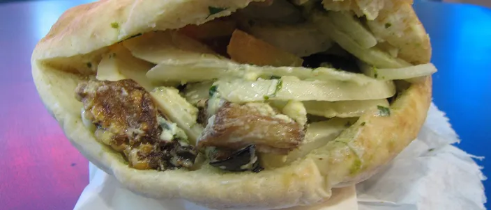 A sabich: a pita split and packed with fried eggplant, egg, sliced potato and chopped salad, standing upright on white napkins on a table.