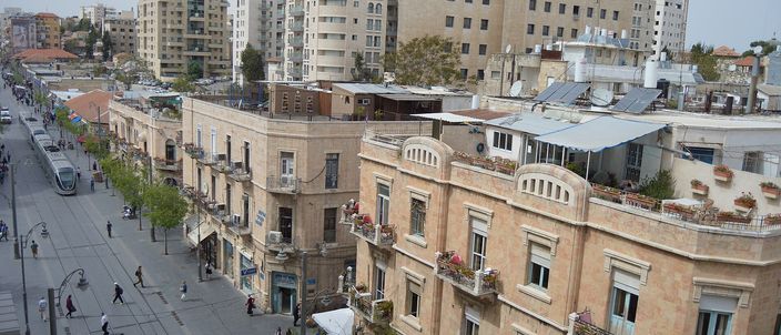 Elevated view down Jaffa Road: a red light-rail tram runs on the pedestrianised street between rows of pale Jerusalem-stone buildings with balconies and rooftop terraces, pedestrians below.