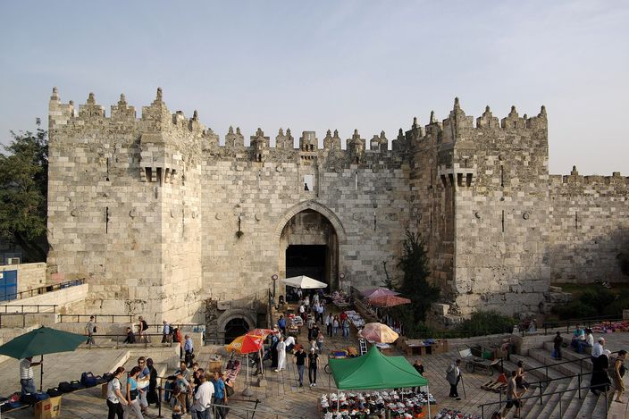 Damascus Gate seen from outside and above: a crenellated twin-towered stone gatehouse with a pointed arched doorway, reached by wide steps descending into a sunken paved plaza where people stand among market umbrellas and stalls.