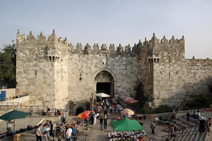 Damascus Gate seen from outside and above: a crenellated twin-towered stone gatehouse with a pointed arched doorway, reached by wide steps descending into a sunken paved plaza where people stand among market umbrellas and stalls.