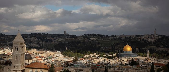 Rooftops of Jerusalem's Old City under a heavy grey sky: the gilded Dome of the Rock on its raised platform at right, a tall white church bell tower at left, tiled and domed roofs between them, and the wooded ridge of the Mount of Olives on the skyline behind.