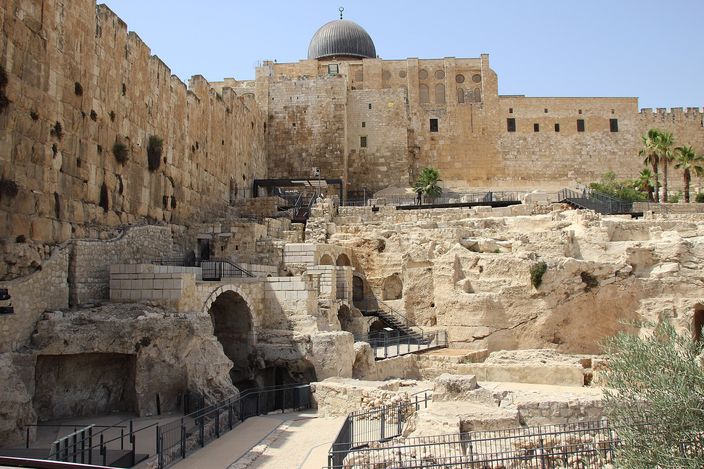 The excavated Jerusalem Archaeological Park below the Temple Mount: a field of exposed stone foundations, vaults and arches with metal walkways among them, the massive Herodian retaining wall rising on the left and the ramparts and lead-grey dome of the Al-Aqsa compound along the skyline above.