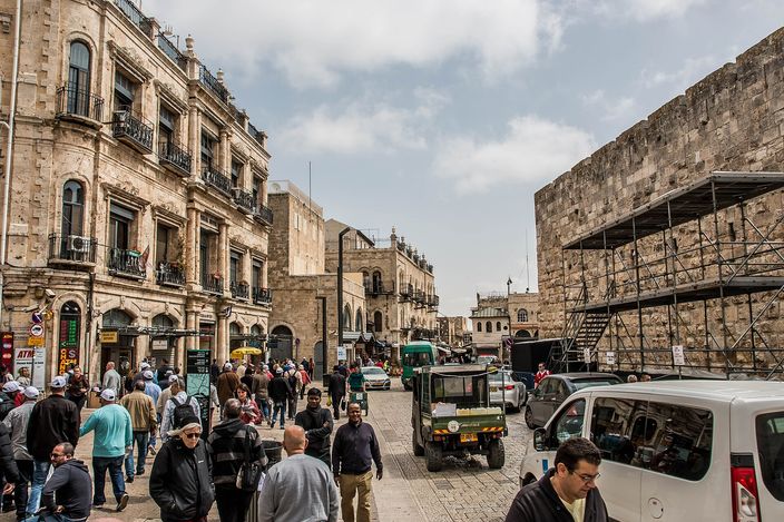 Crowds walking a broad cobbled plaza just inside Jaffa Gate, flanked by honey-coloured stone buildings and the massive Old City wall on the right, with a section under scaffolding.