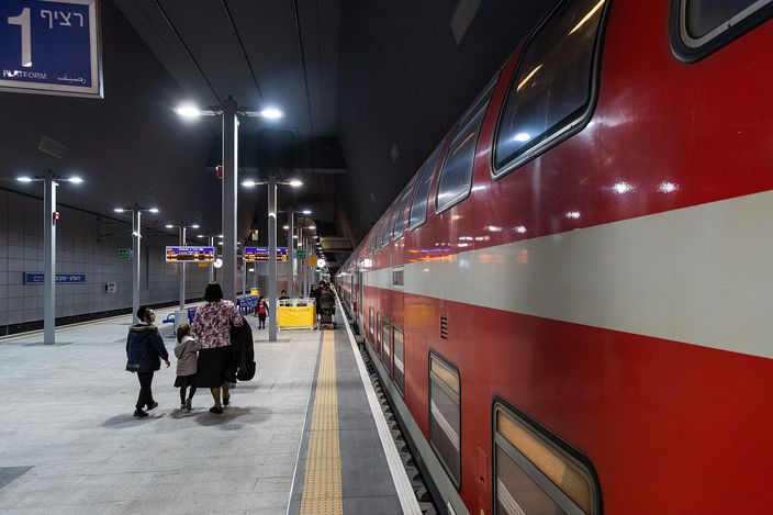 A red-and-white double-deck Israel Railways train stands at an underground platform at Jerusalem's Yitzhak Navon station; a family with children walks along the platform under electronic destination signs reading 'Platform'.