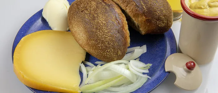 A Halve Hahn laid out on a blue plate: a halved dark rye roll, a thick wedge of yellow Gouda, butter and sliced raw onion, with a pot of brown mustard and a glass of Kölsch beside it