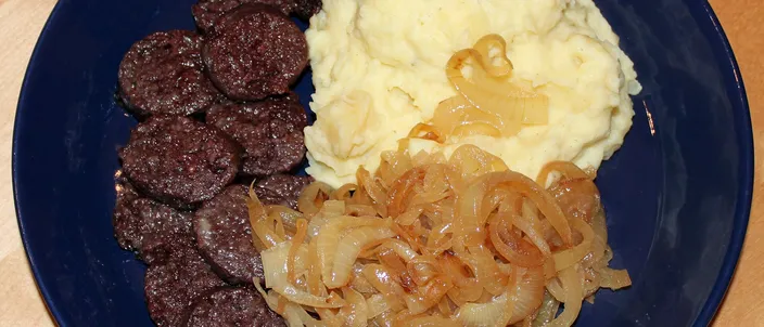 A dark blue plate of Himmel un Ääd: thick slices of fried black pudding beside a mound of mashed potato, topped with a heap of soft fried onions