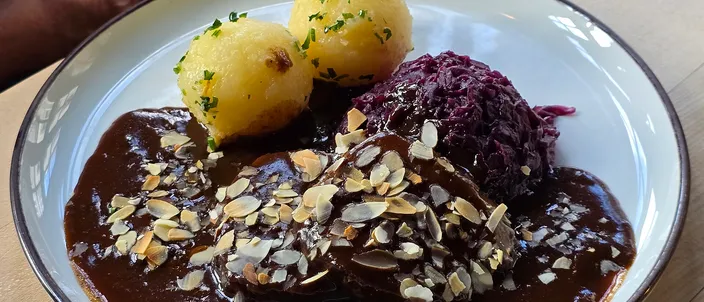A plate of Rhenish sauerbraten: slices of braised beef in a glossy dark brown sweet-sour gravy scattered with flaked almonds, with two potato dumplings and a mound of red cabbage