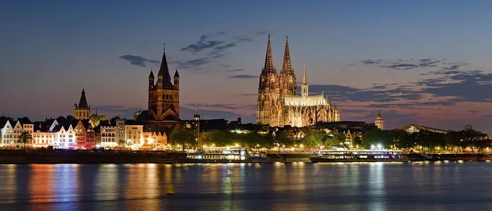 Cologne's old-town riverfront at blue hour seen from across the Rhine, the lit tower of Great St. Martin and the twin spires of the cathedral above a row of narrow gabled houses, their lights reflected in the river