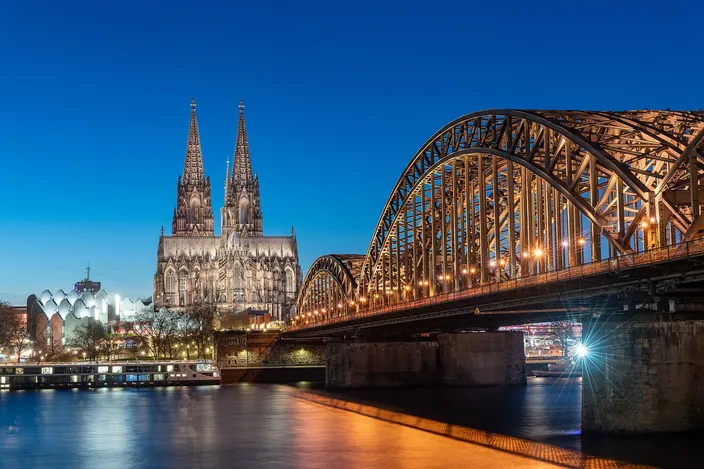 The Hohenzollern Bridge's steel arches lit at blue hour, leading across the Rhine to Cologne Cathedral and the station roof on the far bank