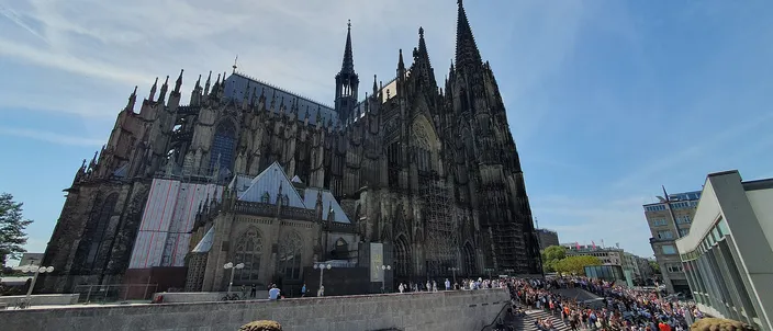 The south flank and twin west towers of Cologne Cathedral rising above the paved cathedral plaza on a bright day, crowds on the steps below