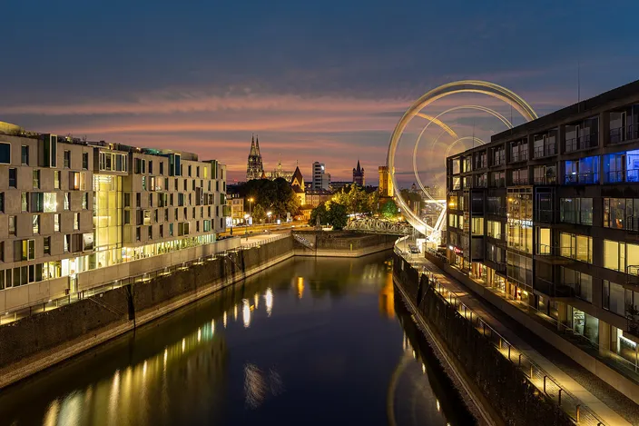 The Rheinauhafen at dusk: modern offices and apartments lining both sides of the old harbor basin, lights reflected in the water, the cathedral's spires on the skyline