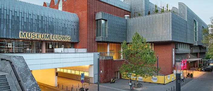 Museum Ludwig at dusk, its zinc-clad roofline and red-brick walls lit from inside above the entrance plaza beside Cologne Cathedral