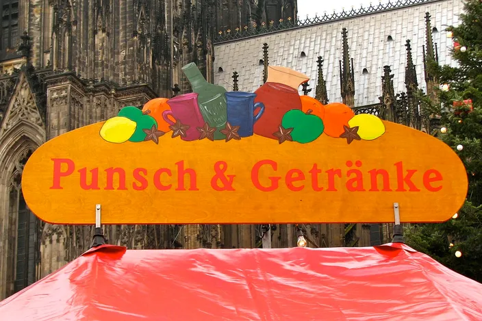 A painted wooden sign reading Punsch und Getränke, decorated with jugs and fruit, over the red roof of a drinks stall, the Gothic stonework of Cologne Cathedral behind it