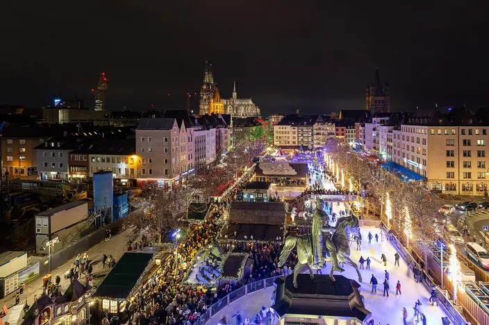 Heumarkt in Cologne at night from above: the Christmas market's lit avenues and ice rink around the square's equestrian statue, the old town's roofs beyond and the cathedral's two spires on the skyline