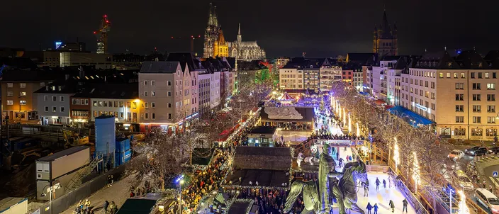 Heumarkt in Cologne at night from above: the Christmas market's lit avenues and ice rink around the square's equestrian statue, the old town's roofs beyond and the cathedral's two spires on the skyline