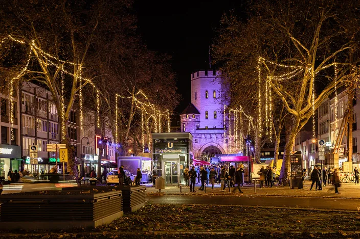 Chlodwigplatz at night in late November, plane trees strung with lights around the Christmas market stalls, and the medieval Severinstorburg gate lit in violet behind them