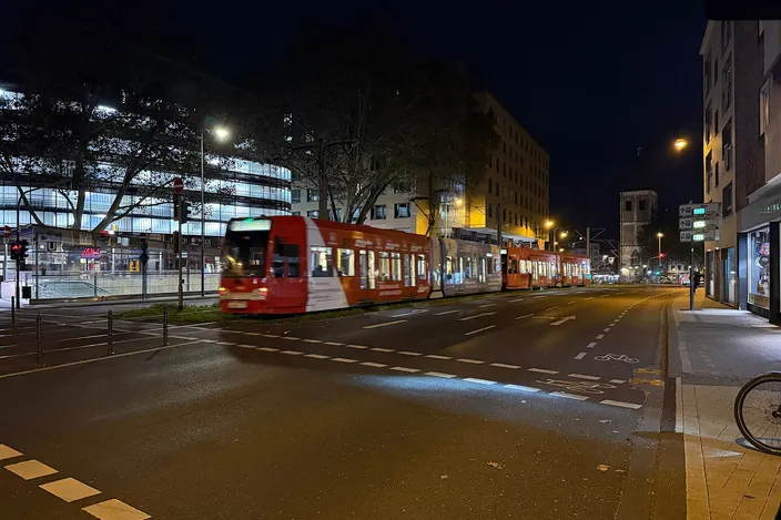 A red KVB low-floor tram on a wide city street at night near Pipinstraße in central Cologne, its interior lit, a church tower at the end of the street
