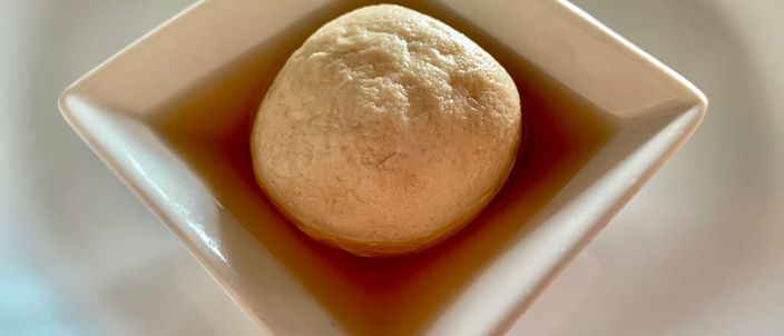 A single pale rosogolla sitting in dark date-palm jaggery syrup in a small white dish.