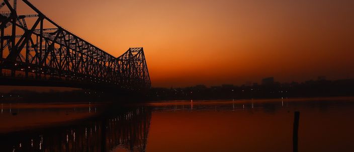 The Howrah Bridge cantilever silhouetted against a deep orange dawn sky, its steel span reflected in the calm Hooghly River with mooring poles in the foreground.