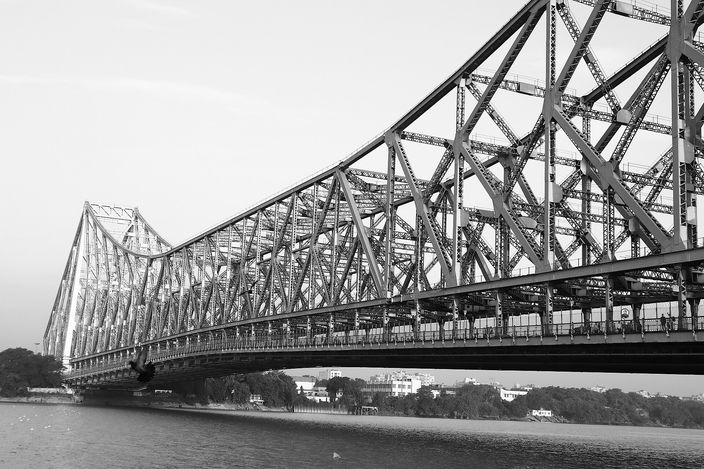 The riveted steel cantilever of the Howrah Bridge in monochrome, seen low from the Kolkata bank, its trusswork receding across the Hooghly toward the far shore.