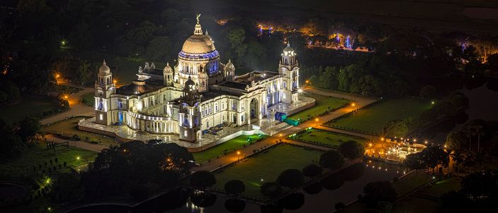 The floodlit white marble dome and colonnaded wings of the Victoria Memorial seen from above at night, ringed by lamplit garden paths, clipped hedges and a dark reflecting pool.