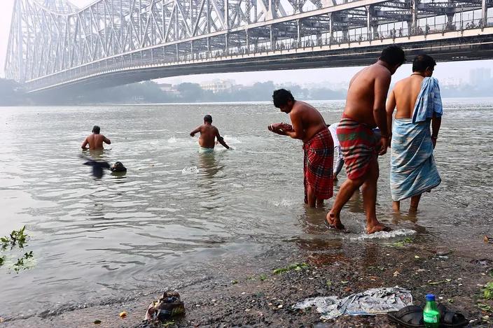 Men bathing and washing at a muddy bank of the Hooghly, waist deep in brown water, with the steel lattice of the Howrah Bridge crossing the hazy sky above them.