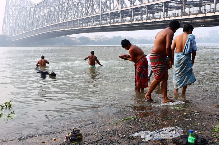 Men bathing and washing at a muddy bank of the Hooghly, waist deep in brown water, with the steel lattice of the Howrah Bridge crossing the hazy sky above them.