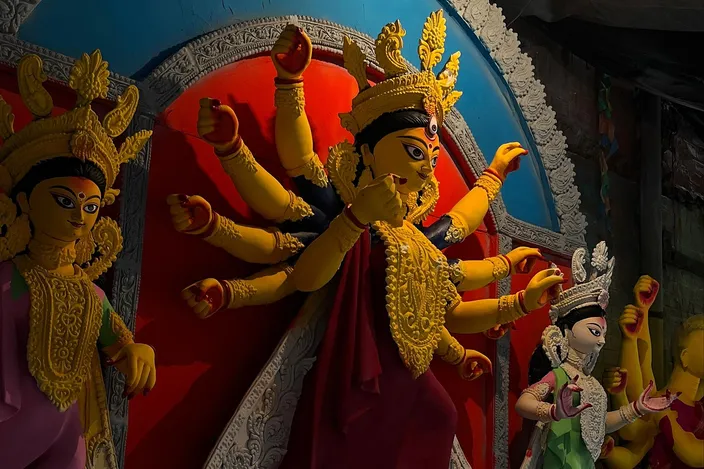 A finished clay Durga idol painted yellow and crimson, her many arms fanned out around a gilded bodice, flanked by smaller painted deities and a roaring lion, in the dim interior of an idol-maker's workshop.