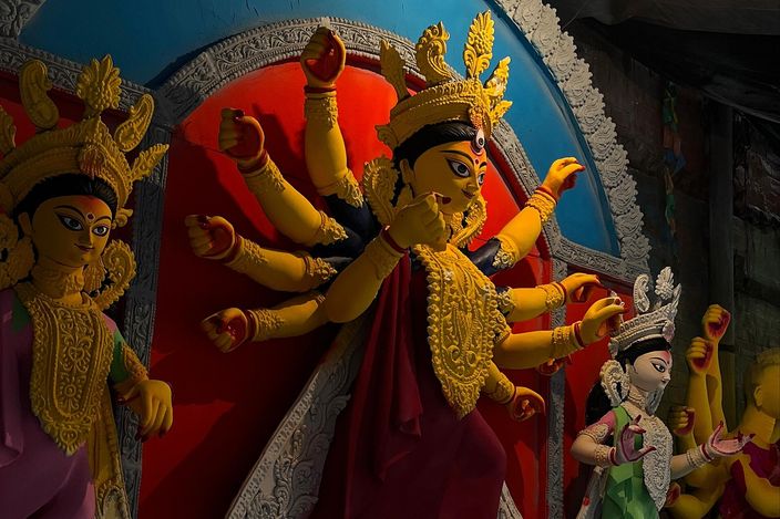 A finished clay Durga idol painted yellow and crimson, her many arms fanned out around a gilded bodice, flanked by smaller painted deities and a roaring lion, in the dim interior of an idol-maker's workshop.