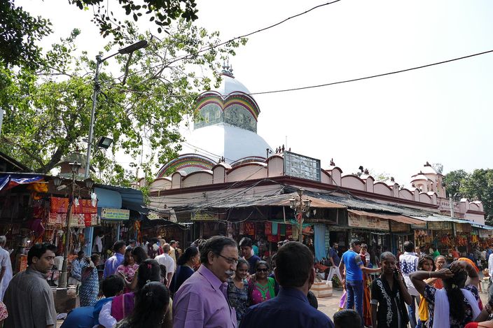 A crowd of visitors filling a lane in front of the Kalighat Kali temple, its pink arcaded wall and white ribbed dome rising behind rows of awninged stalls hung with garlands and trinkets.