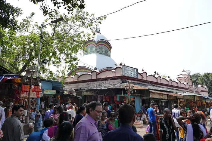 A crowd of visitors filling a lane in front of the Kalighat Kali temple, its pink arcaded wall and white ribbed dome rising behind rows of awninged stalls hung with garlands and trinkets.