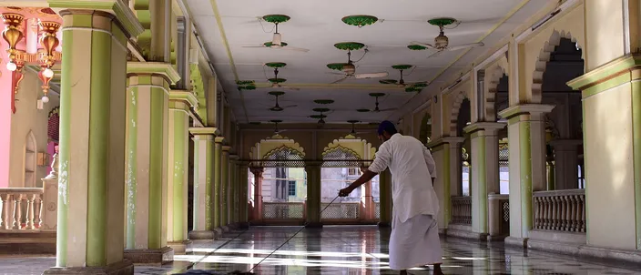 A long arcaded veranda inside a mosque, pale green columns and pointed arches receding into shadow, a man in a white kurta sweeping the polished checkerboard floor where a band of daylight falls across it.