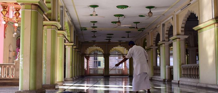 A long arcaded veranda inside a mosque, pale green columns and pointed arches receding into shadow, a man in a white kurta sweeping the polished checkerboard floor where a band of daylight falls across it.