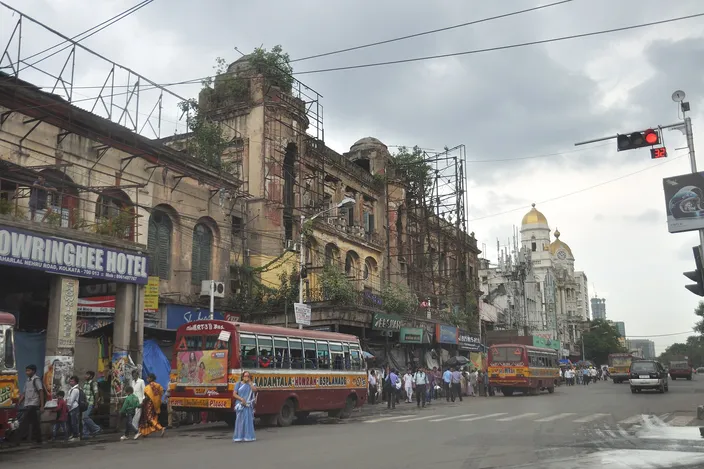 A crossing at Esplanade, Chowringhee Road, with the crumbling scaffolded facade of the old Chowringhee Hotel, purple and yellow public buses lined up, and the domed Metropolitan Building visible under a gray monsoon sky