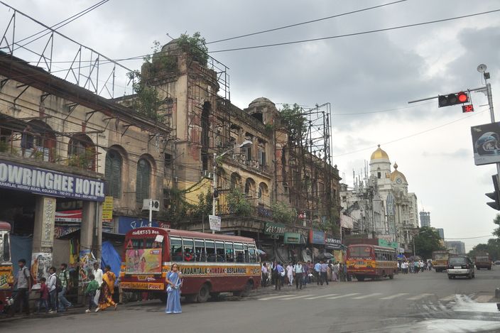 A crossing at Esplanade, Chowringhee Road, with the crumbling scaffolded facade of the old Chowringhee Hotel, purple and yellow public buses lined up, and the domed Metropolitan Building visible under a grey monsoon sky