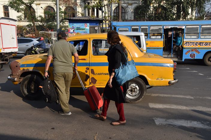 Two travellers with a wheeled suitcase board a battered yellow Ambassador taxi at a busy Kolkata crossing, a blue-and-yellow city bus behind them