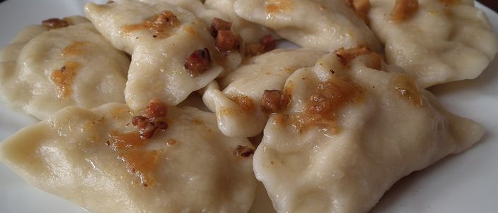 A white plate of plump boiled half-moon dumplings, glossy with melted butter and scattered with crisp golden bits of fried onion and bacon.