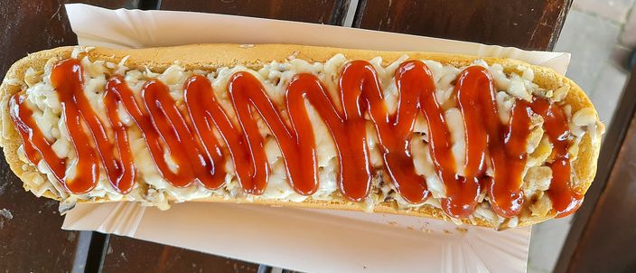 A long half-baguette open-face, toasted, covered in melted cheese and mushrooms with a zig-zag of ketchup down its length, resting on paper.