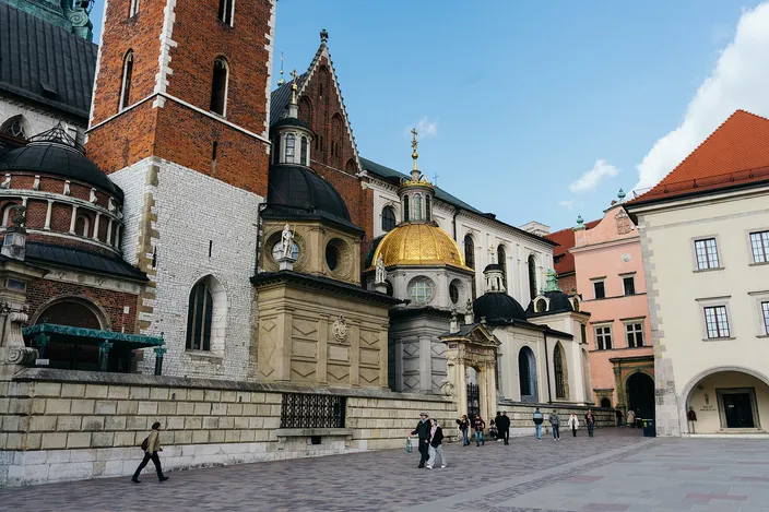 The cathedral square on Wawel hill: a tall brick tower and a Gothic gable on the left, a gilded dome and two dark cupolas over the chapels in the center, a pink and a white building on the right, and a dozen people crossing the paved square.