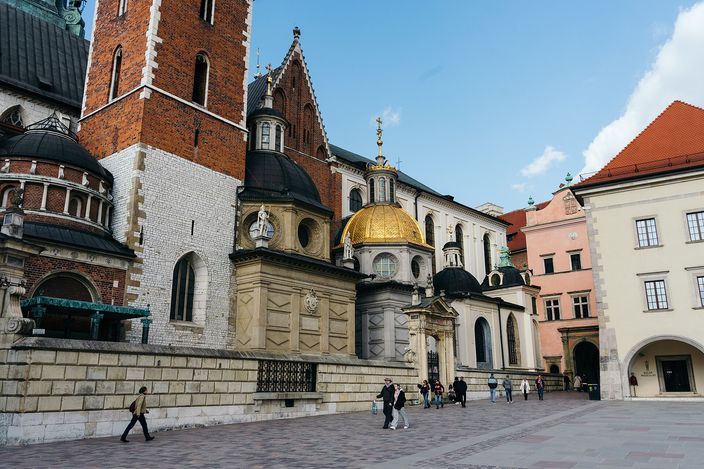 The cathedral square on Wawel hill: a tall brick tower and a Gothic gable on the left, a gilded dome and two dark cupolas over the chapels in the centre, a pink and a white building on the right, and a dozen people crossing the paved square.