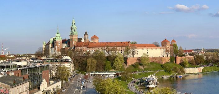 Wawel hill seen from across the Vistula: the long red-roofed castle and the green spires of its cathedral standing on a brick-walled rock, with the tree-lined embankment, a road and moored river boats along the water below.