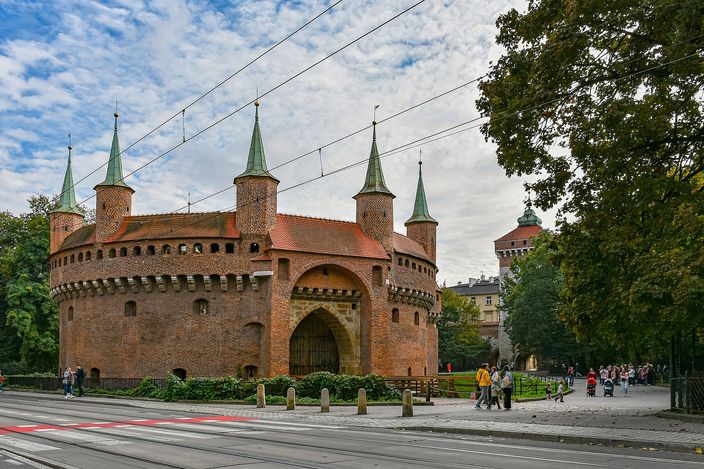 The Kraków Barbican, a circular red-brick fortress with seven green-spired turrets and a pointed gate arch, autumn trees and strolling passers-by along the Planty behind it, tram wires strung overhead and tram rails in the road in front.