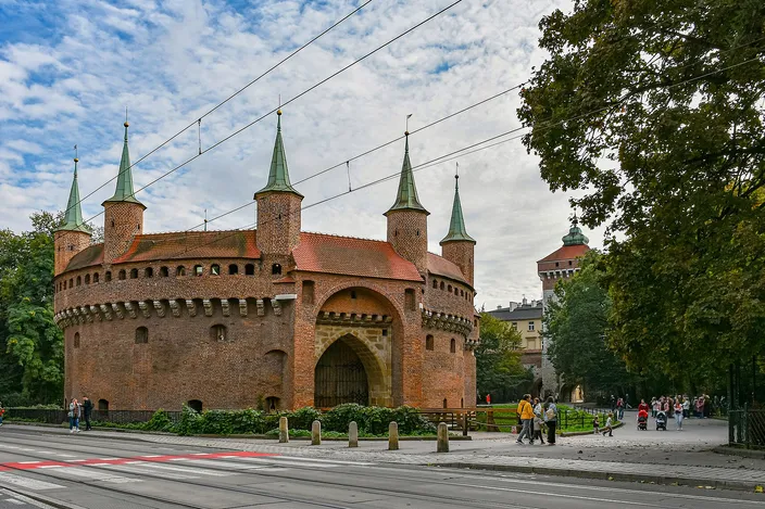 The Kraków Barbican, a circular red-brick fortress with seven green-spired turrets and a pointed gate arch, autumn trees and strolling passers-by along the Planty behind it, tram wires strung overhead and tram rails in the road in front.