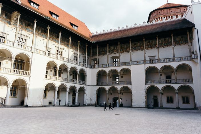 The three-tier Renaissance arcaded courtyard of Wawel Castle, slender columns and a painted frieze running the length of two wings under a red tiled roof, with two people crossing the empty flagstones.