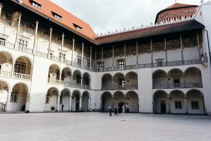 The three-tier Renaissance arcaded courtyard of Wawel Castle, slender columns and a painted frieze running the length of two wings under a red tiled roof, with two people crossing the empty flagstones.