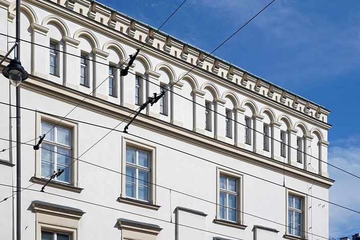 The upper two storeys of a white Kazimierz facade under a deep blue sky, a row of small blind arches running below the cornice and tall shuttered windows beneath, with tram wires and a streetlamp crossing the frame.
